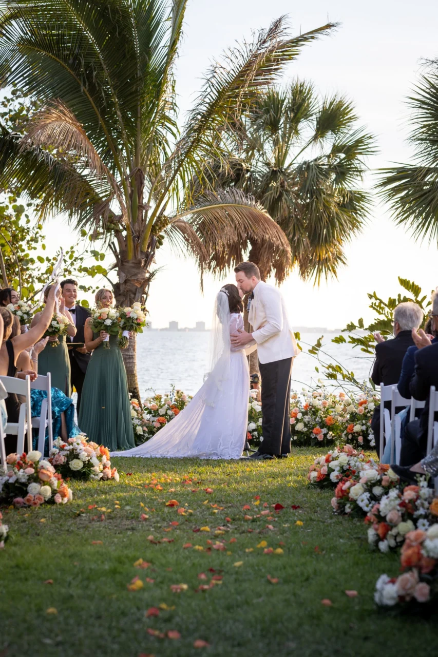 Brud og brudgom kysser under utendørs vielse ved sjøen omgitt av palmer og blomsteroppsatser