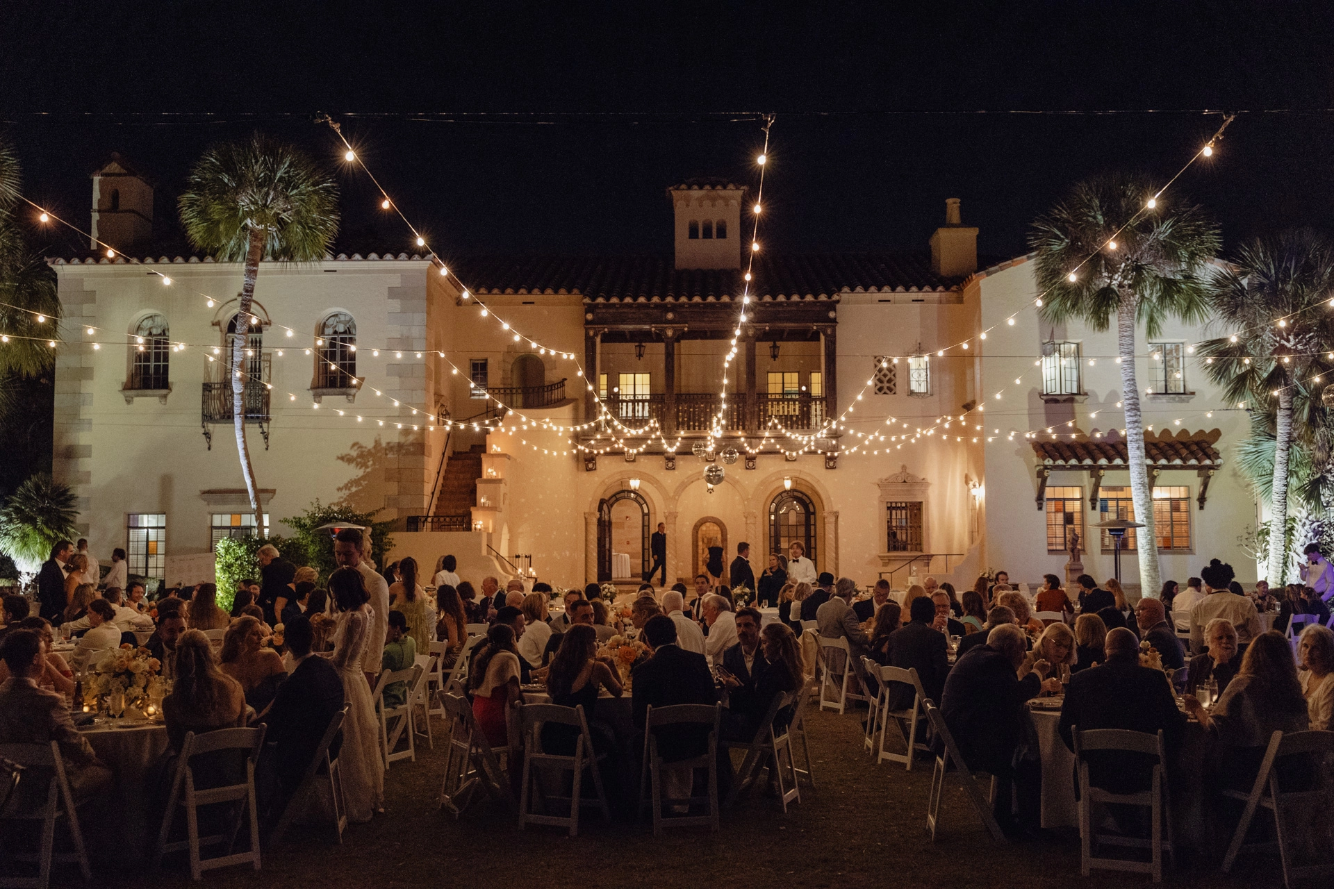 Stemningsfull bryllupsmiddag utendørs med lysslynger foran historisk bygning om kvelden